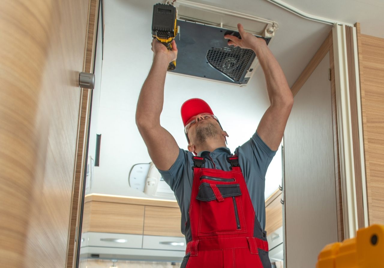Frustrated RV owner inside a hot camper with fan blowing, looking at a phone for help